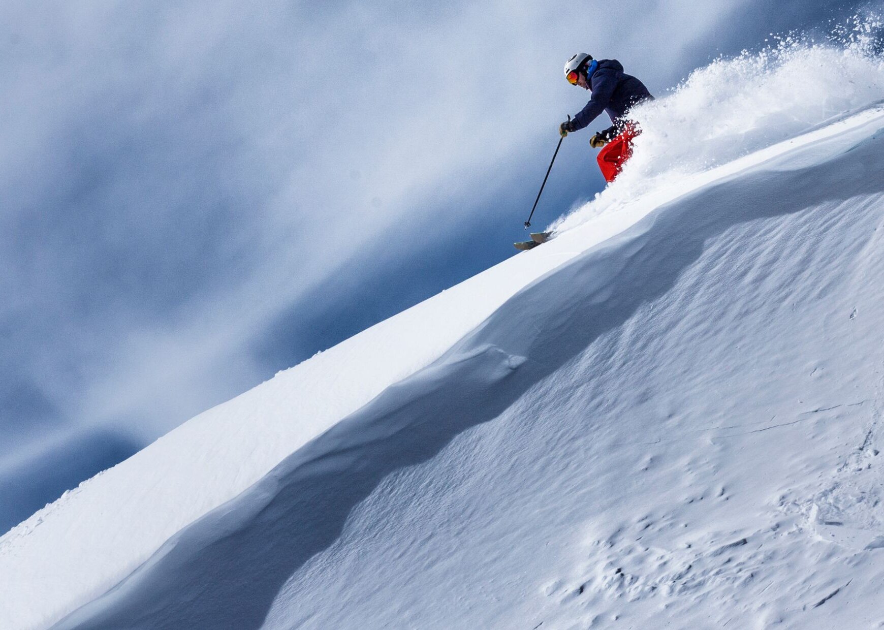 A freerider in red pants rides down a steep snow-covered slope, kicking up powder snow.